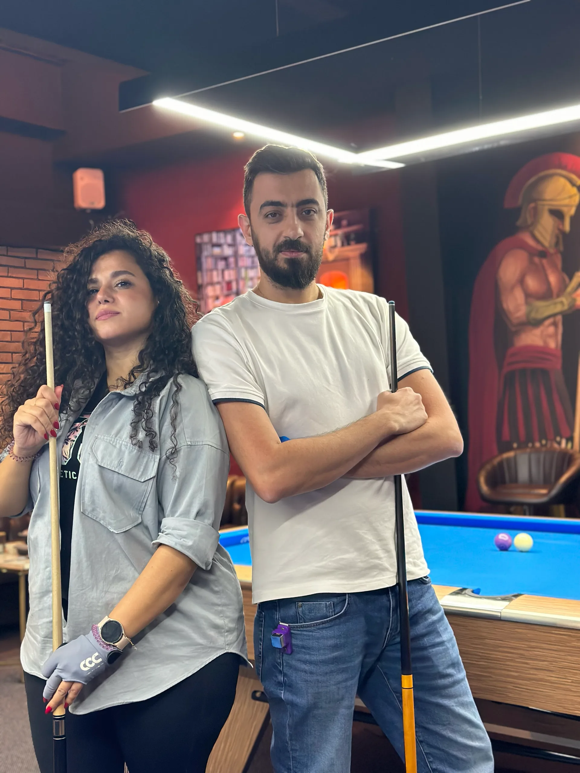 Two players standing with their cues beside a billiard table in front of a Spartan mural