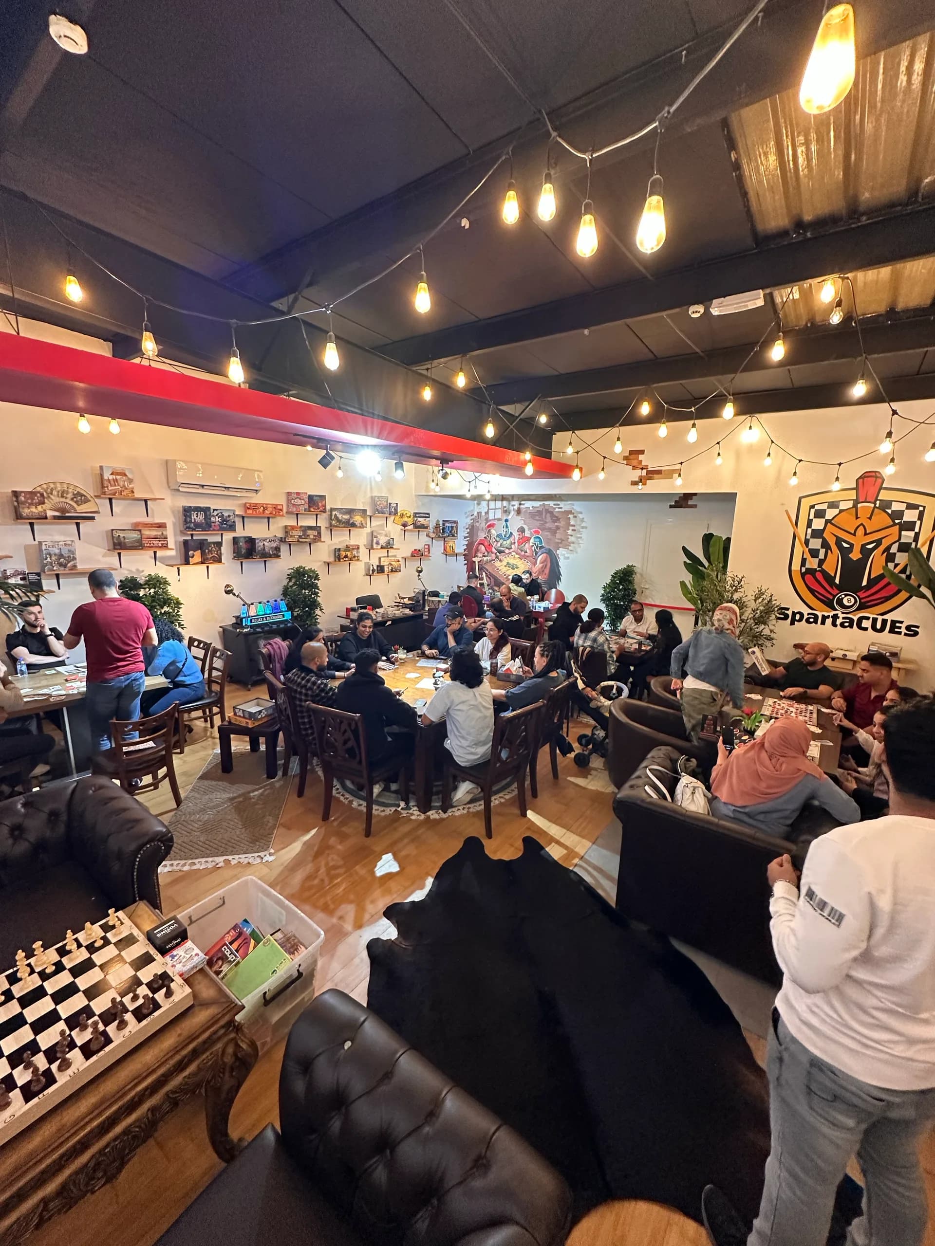 The board game room at Spartacues with every table taken, under strings of warm lights