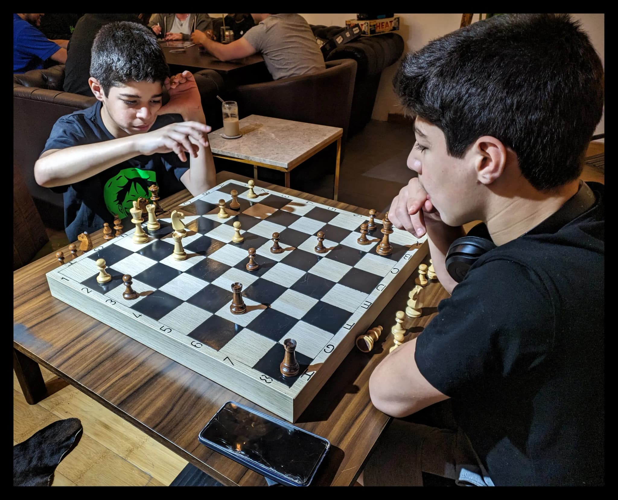 Two players facing each other over a chess board, with other tables busy behind them
